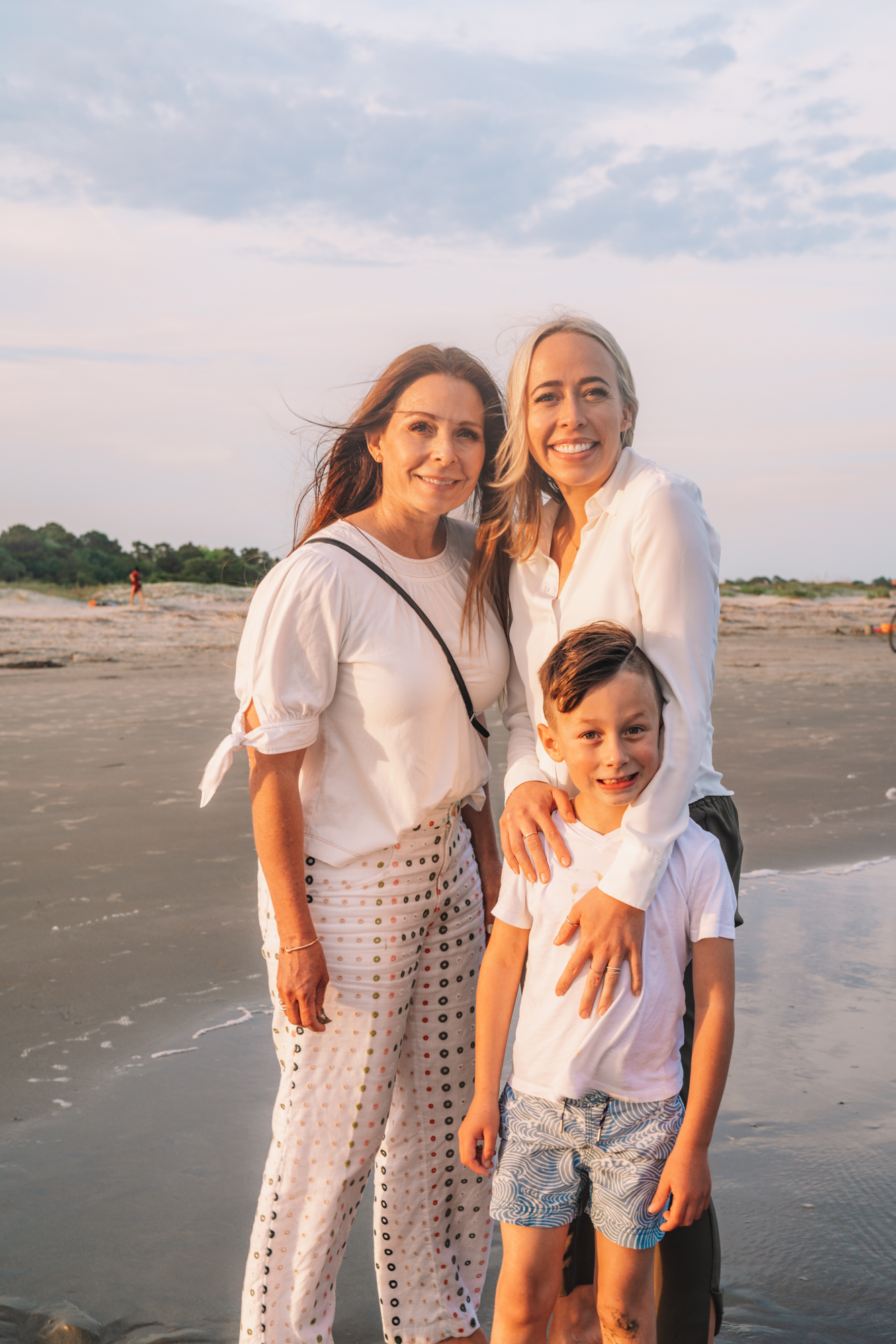 a small family smiles on the shore of the beach during sunset.